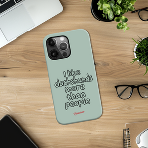 Saucisse sage-green phone case with “I like dachshunds more than people” lettering and a red signature, on a wooden desk.