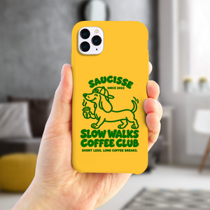 Saucisse yellow case with green dachshund artwork and Slow Walks Coffee Club lettering, held in a hand in a home setting.