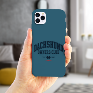 Saucisse teal case with navy Dachshund Owners Club lettering and a red Saucisse signature, held in a hand in a home setting.