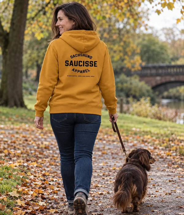 Woman wearing the Curcuma Saucisse Manchester England hoodie, showing its back print while walking a long-haired dachshund in an autumn park.