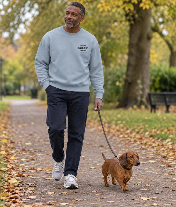 Man wearing the Saucisse Manchester England unisex sweater in Aquamarine while walking a dachshund in a leafy park