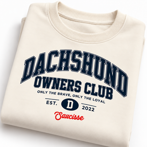 Folded Ivory Saucisse Dachshund Owners Club 2026 unisex sweater showing the navy varsity print and red Saucisse signature