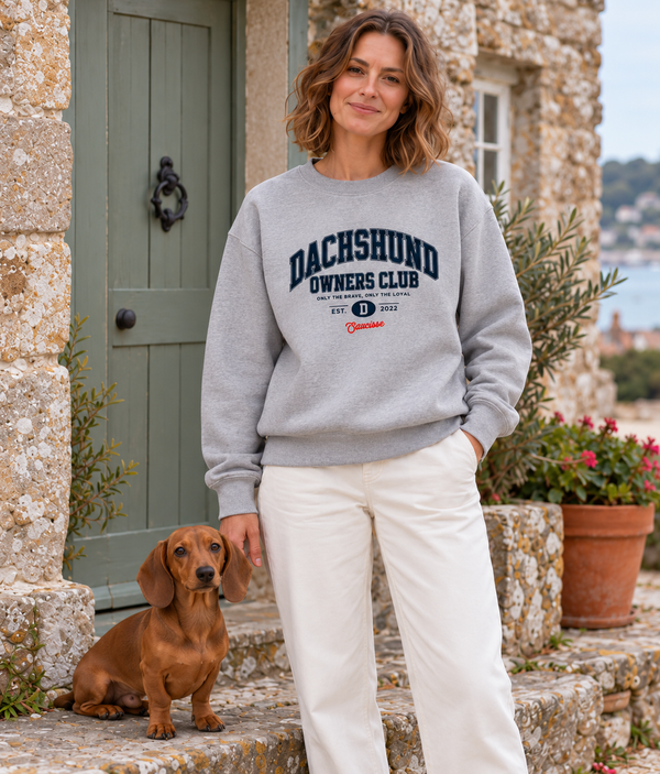 Woman wearing the Saucisse Dachshund Owners Club 2026 unisex sweater in Moon Heather Grey beside a small smooth-haired red dachshund outside a stone cottage