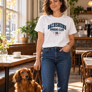 Woman wearing the white Saucisse Dachshund Owners Club 2026 unisex T-shirt at a cafe with a long-haired dachshund.