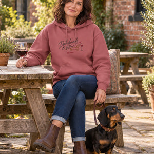 A woman wearing the Antique Rose Saucisse Dachshunds & Wine hoodie in a British pub garden, with a dachshund at her feet.