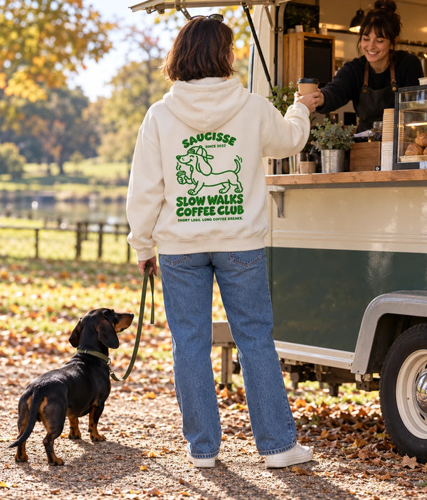 Saucisse Slow Walks Coffee Club unisex hoodie in Ivory, rear coffee-stop lifestyle view with green back print and a dachshund on a lead