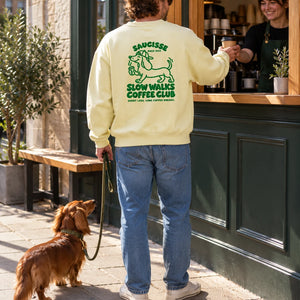 Saucisse Slow Walks Coffee Club unisex sweater in Lemon Citrus, rear coffee-stop lifestyle view with green back print and a dachshund on a lead