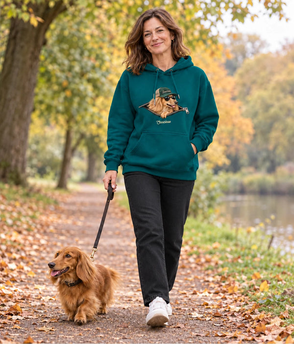 Woman wearing the Saucisse Unzipped long-haired unisex hoodie in Peacock Green while walking a long-haired dachshund beside the water.