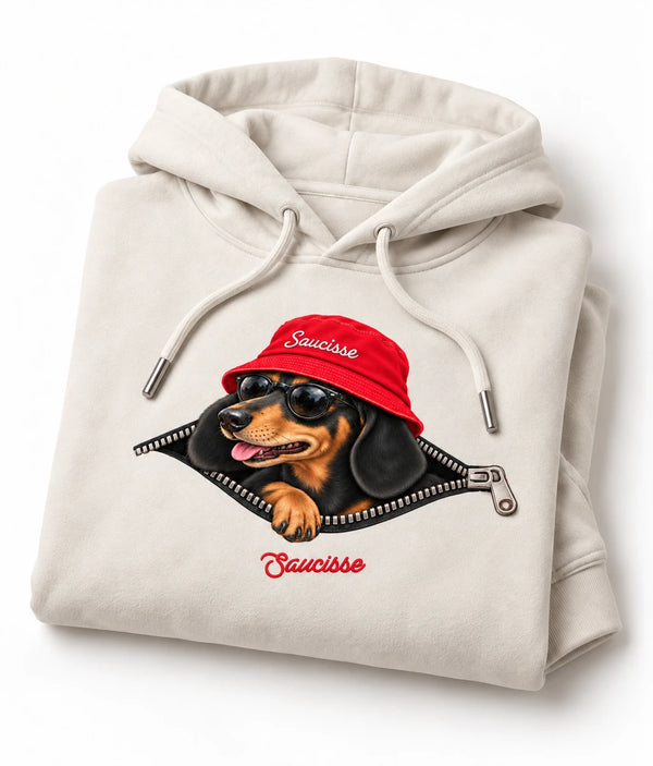 Folded Ivory Saucisse Unzipped Short Haired unisex hoodie on a white studio background, showing a dachshund in a red Saucisse bucket hat and sunglasses peeping through an illustrated zip and the red signature.