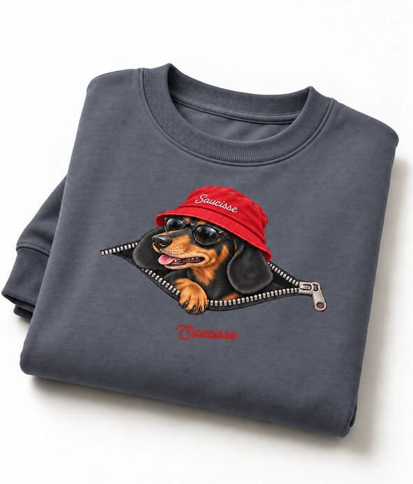 Folded Mineral Grey Saucisse Unzipped Short Haired unisex sweater on a white studio background, showing a dachshund in a red Saucisse bucket hat and sunglasses peeping through an illustrated zip and the red signature.