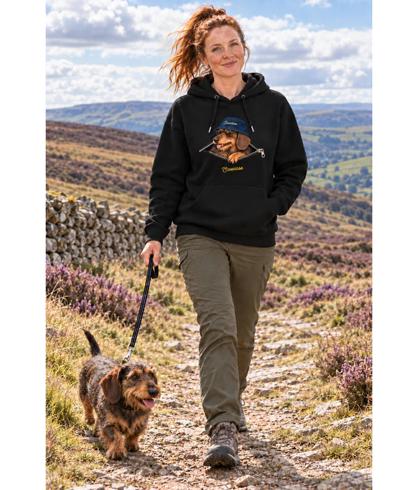 Woman wearing the Saucisse Unzipped wire-haired unisex hoodie in Black while walking a wire-haired dachshund along an open moorland path.