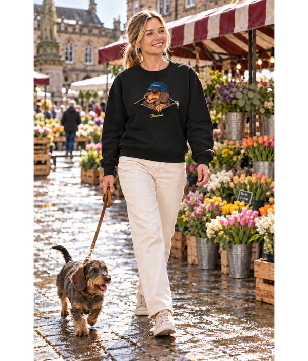 Woman wearing the Saucisse Unzipped wire-haired unisex sweater in Black while walking a wire-haired dachshund through a flower market.