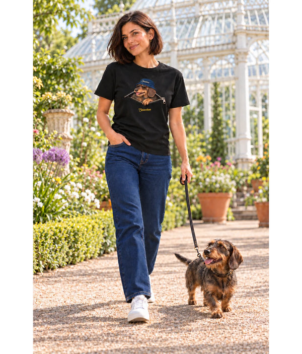 Woman wearing the Saucisse Unzipped wire-haired unisex T-shirt in Black while walking a wire-haired dachshund beside a garden glasshouse.