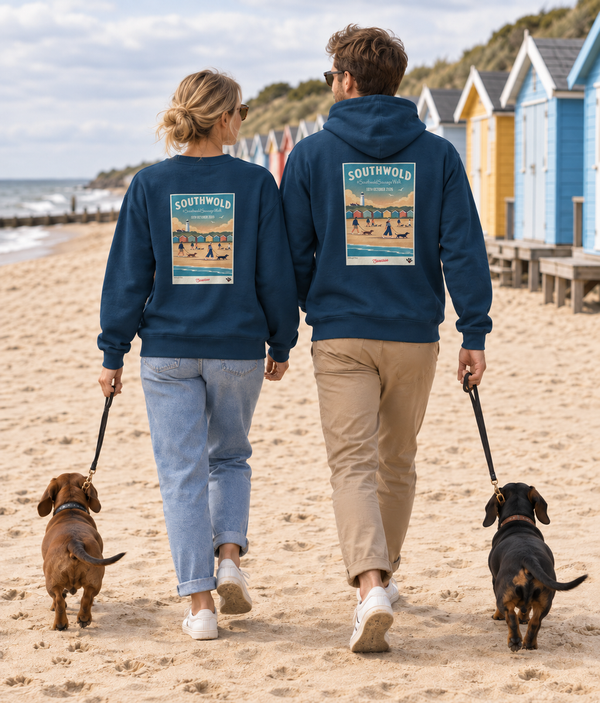 Couple wearing Southwold Sausage Dog Walk 2026 Saucisse hoodie and sweater while walking dachshunds by beach huts