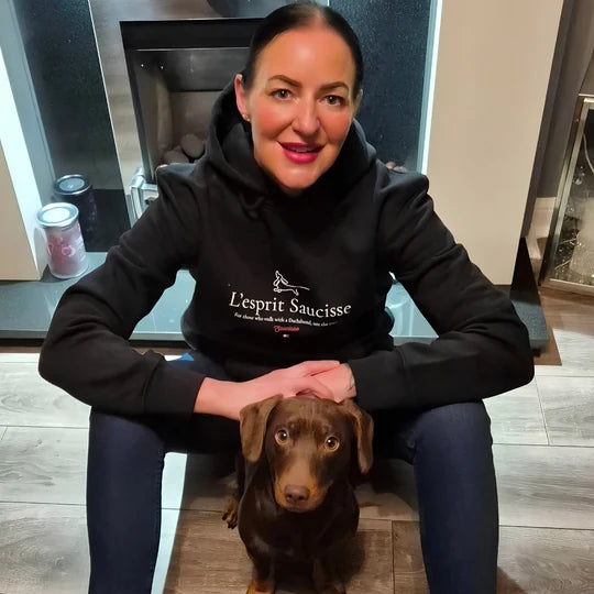 A Saucisse customer in a black L’esprit Saucisse hoodie with a chocolate dachshund at home