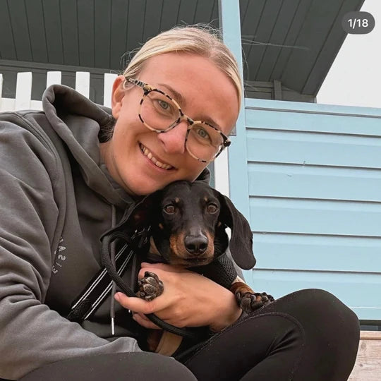 A customer wearing a grey Saucisse hoodie cuddling a dachshund beside a beach hut