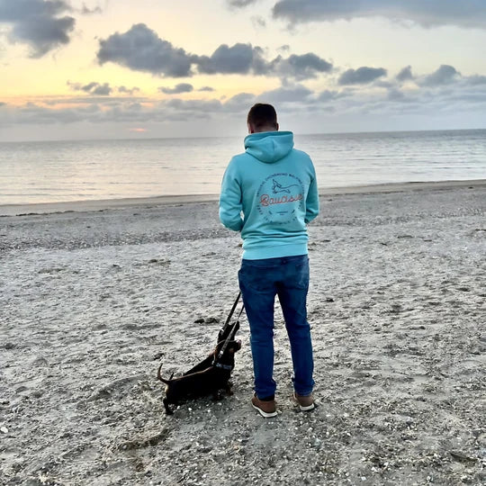 A customer in a blue Saucisse hoodie walking a dachshund on the beach at sunset